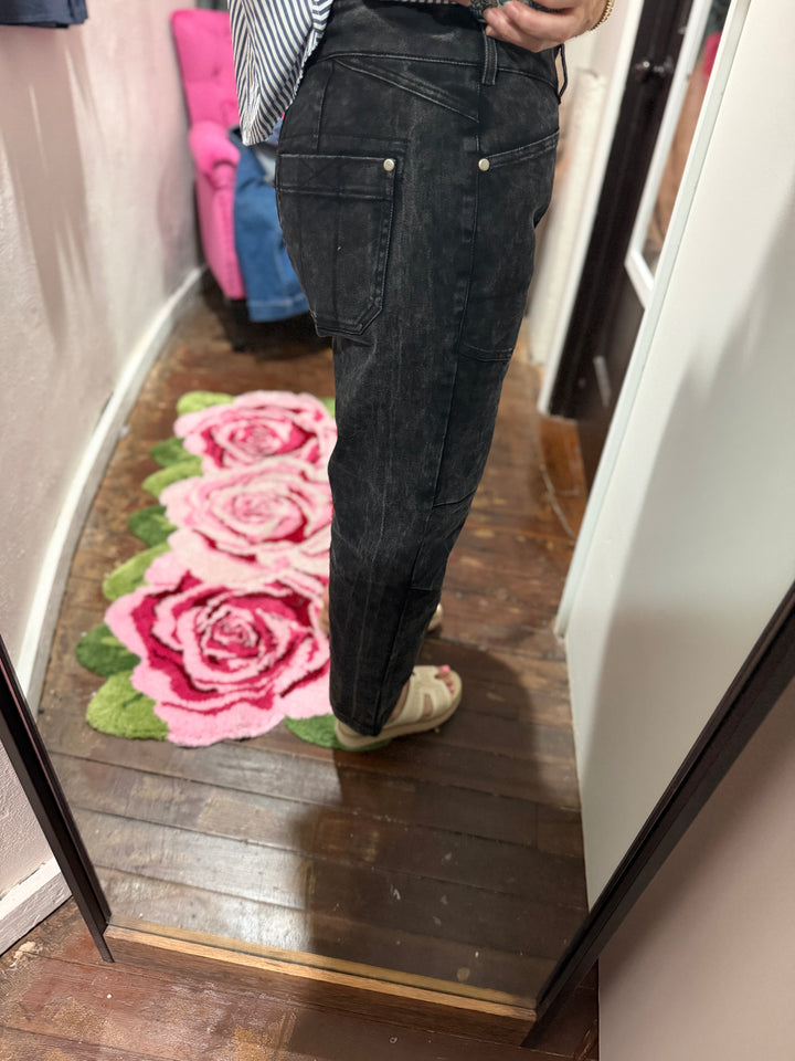 A person wearing dark washed denim jeans and beige slip-on shoes, standing in a mirror to show the fit of the jeans in a home setting with a wooden floor and a pink rose rug.