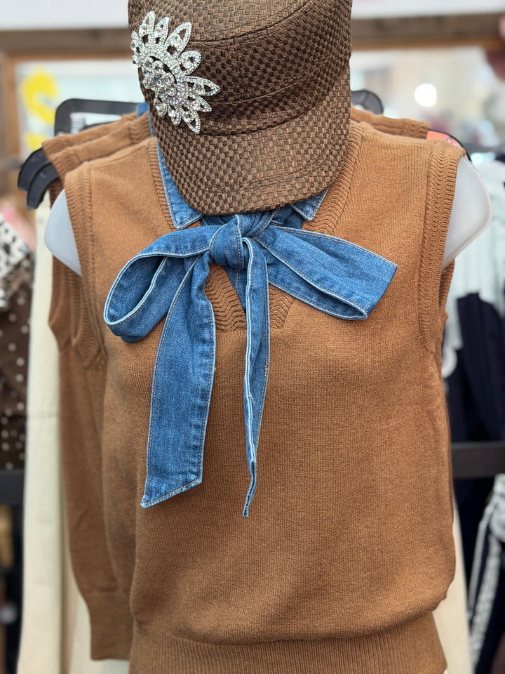 A brown knit sweater vest displayed on a mannequin torso, styled with a large blue denim bow at the neckline and a brown woven hat with a rhinestone brooch.