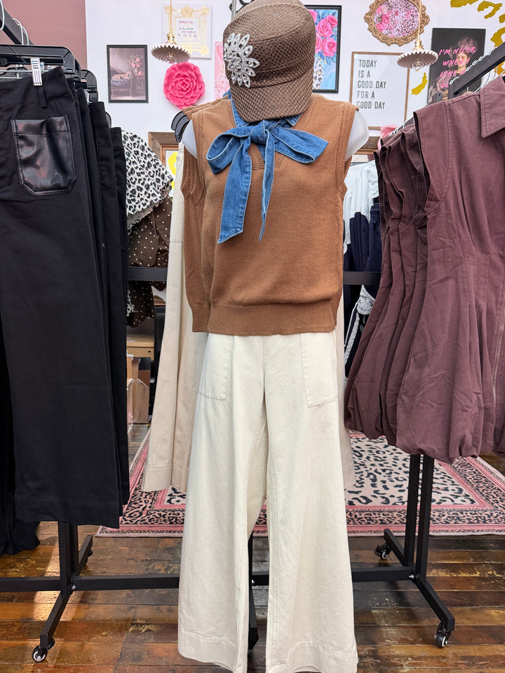 A clothing display mannequin in a boutique setting wearing a brown knit sleeveless top with a large blue denim bow, cream wide-leg pants, a brown textured hat with a white floral embellishment, and a burgundy sleeveless dress hanging on a nearby rack.