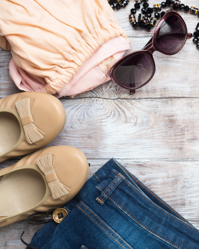 Tan shoes, pink top, blue jeans, and sunglasses on a wooden surface